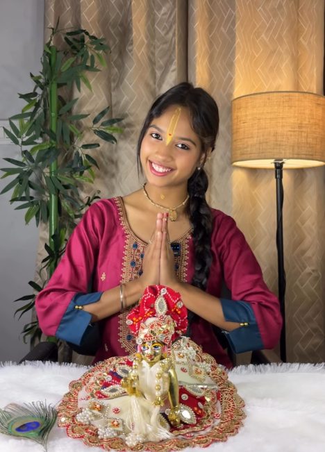 Woman offering namaste beside Laddu Gopal idol wearing a red and white embroidered poshak
